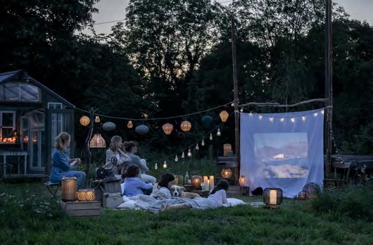 Familie sitzt auf Decken im Garten vor einer projizierten Leinwand zwischen den Bäumen, Lampions, Lichterketten, Dämmerlicht