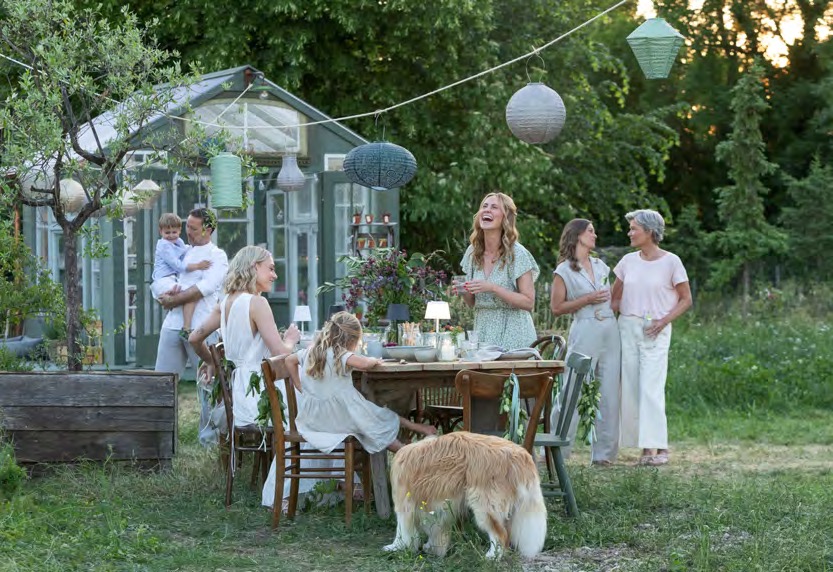 Familie an gedeckter Tafel unter Lampions vor einem mintgrünen Gewächshaus am Abend